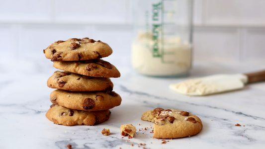 A close-up image featuring a stack of Sourdough Chocolate Chip Cookies, accompanied by a single cookie with a bite taken out. In the background, a jar of 'You Knead Sourdough' starter sits beside a spatula coated in sourdough starter.